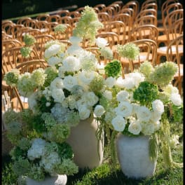 White rose and hydrangea arrangements in stone vases at outdoor wedding seating