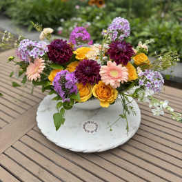 Mixed bouquet of yellow roses, purple blooms, and peach daisies in a white bowl