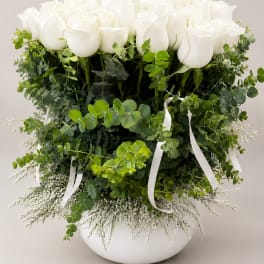 White roses arranged in a round white vase with lush greenery and ribbon accents