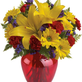 Bright bouquet of yellow lilies, daisies, and red carnations in a red glass vase