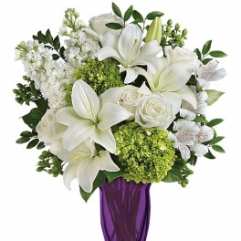 Tall arrangement of white lilies, roses, and green hydrangeas in a purple glass vase