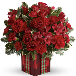 Red roses and carnations in a red cube vase with plaid ribbon and holiday evergreens