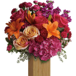 Bright arrangement of orange lilies, pink and peach roses, and magenta blooms in a tall wooden vase