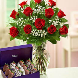 Red rose bouquet in a glass vase beside a purple box of chocolate-covered strawberries