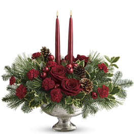 Low red rose and carnation centerpiece with evergreens and two red taper candles in a silver pedestal bowl