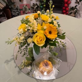 Yellow and white flower arrangement in a textured vase with a pastry container in front