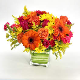 Bright arrangement of orange gerbera daisies, roses, and yellow daisies in a square glass vase with green lining.