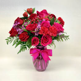 Red and hot pink roses and carnations with purple filler flowers in a pink vase with a bow.