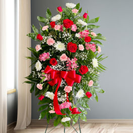 Standing floral spray with red roses, pink carnations, and white daisies on a red ribboned easel