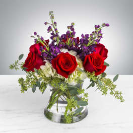 Low arrangement of red roses, purple stock, and white hydrangeas in a clear glass vase with trailing greenery