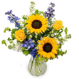 Bouquet of sunflowers, yellow roses, and blue flowers in a clear glass vase