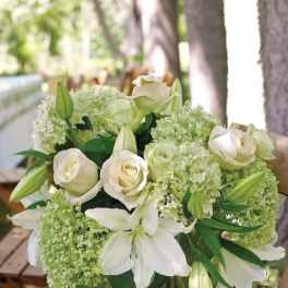 White lilies, roses, and green hydrangeas in a clear glass vase on a wooden stool outdoors