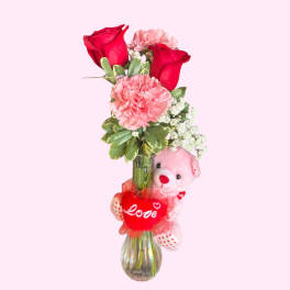Red roses and pink carnations in a glass vase with a teddy bear