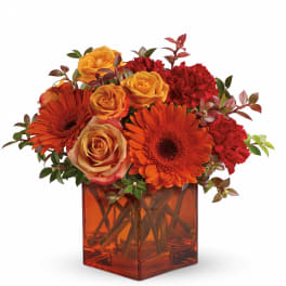 Orange and yellow roses, orange gerbera daisies, and red carnations in an orange glass cube vase