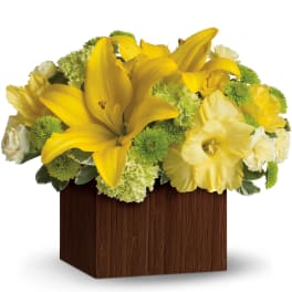 Low arrangement of yellow lilies and mixed yellow, green, and white flowers in a brown wood box