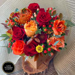 Red and orange rose arrangement with protea and yellow mums in a wooden box on a wood slice base