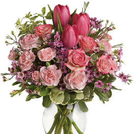 Pink tulips, roses, and carnations with small purple flowers arranged in a clear glass vase