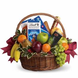 Wicker gift basket filled with assorted fruits and chocolate bars, accented with holly and red bows.