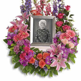 Colorful circular sympathy wreath of pink lilies, roses, and gerberas framing a silver photo frame.