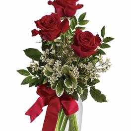 Three red roses with small white flowers in a clear glass vase with a red ribbon bow