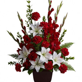 Tall arrangement of red carnations, red gladiolus, and white lilies in a dark pedestal vase