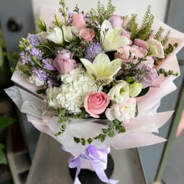 Pastel bouquet with pink roses, white lilies, and lavender flowers wrapped in pale paper