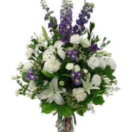 Tall arrangement of white lilies and carnations with purple delphinium and iris in a clear glass vase