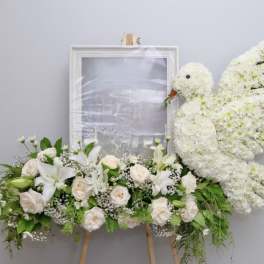 White floral funeral tribute with roses, lilies, and a dove-shaped spray on an easel