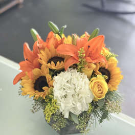 Orange lilies, sunflowers, a white hydrangea, and yellow roses in a low black container.