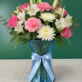 Pink roses, carnations, and white daisies in a blue glass vase with a ribbon