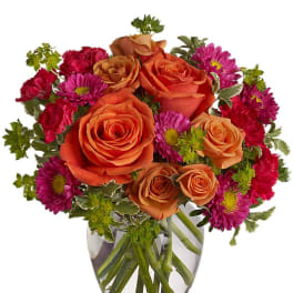 Compact arrangement of orange roses with bright pink mums and carnations in a clear glass vase