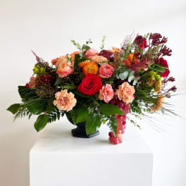 Mixed bouquet with red roses, peach carnations, and orange blooms in a black vase