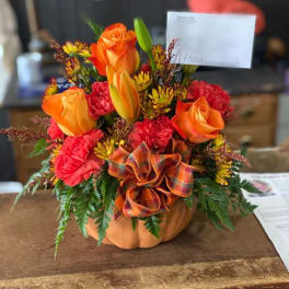 Orange and red floral arrangement in a pumpkin container with a plaid bow