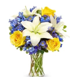Bouquet of white lilies, yellow roses, and blue flowers in a clear glass vase