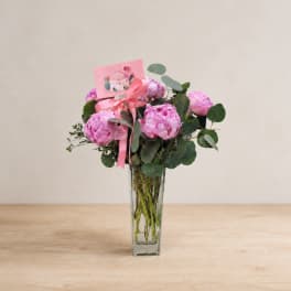 Pink peony bouquet in a clear glass vase with a ribbon and card