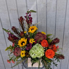 Mixed bouquet with sunflowers, orange roses, hydrangea, and burgundy blooms in a wooden container.