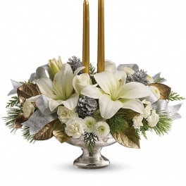 Low centerpiece of white lilies and other white flowers with two gold taper candles in a silver bowl.