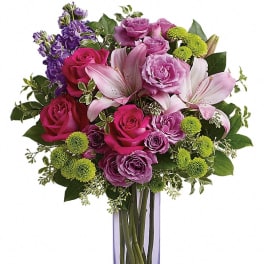 Tall arrangement of pink roses, lavender blooms, lilies, and green button mums in a purple glass vase