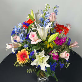 Mixed bouquet with lilies, roses, gerbera daisies, and blue flowers in a glass vase