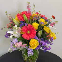 Colorful mixed bouquet in a clear glass vase with gerbera daisies, roses, lilies, and purple filler flowers