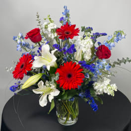 Mixed bouquet with red gerbera daisies, red roses, white lilies, and blue flowers in a glass vase