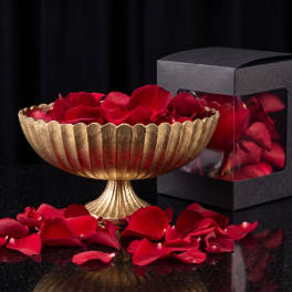 Gold pedestal bowl filled with red rose petals beside a black gift box
