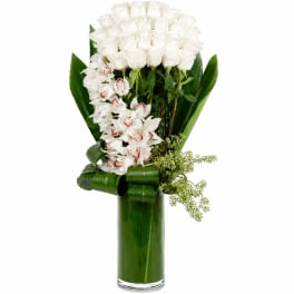 White roses and orchids in a tall glass vase with broad green leaves