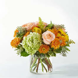 Mixed bouquet of peach roses, green hydrangea, and orange mums in a glass vase