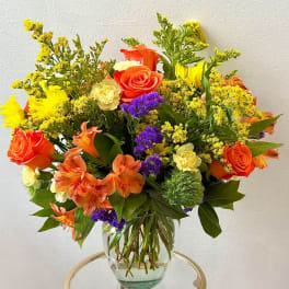 Mixed bouquet of orange roses, yellow blooms, and purple flowers in a glass vase