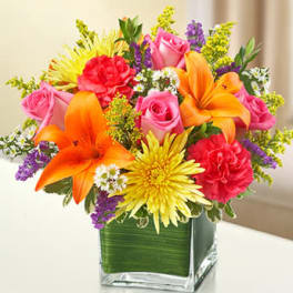 Bright mixed bouquet with orange lilies, pink roses, and yellow mums in a green-lined square glass vase.
