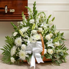 White lilies, roses, and mums in a sympathy basket with a white bow beside a wooden casket.