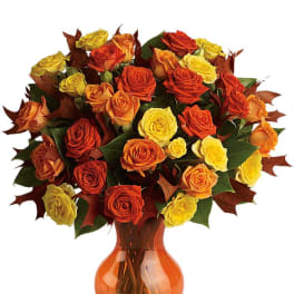Arrangement of orange and yellow roses with fall leaves in an orange glass vase