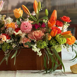 Low wooden box centerpiece filled with orange, pink, and peach blooms including roses, tulips, and lilies.