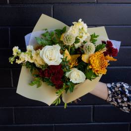 Hand-tied bouquet of white roses, yellow chrysanthemums, and dark red blooms wrapped in pale paper.
